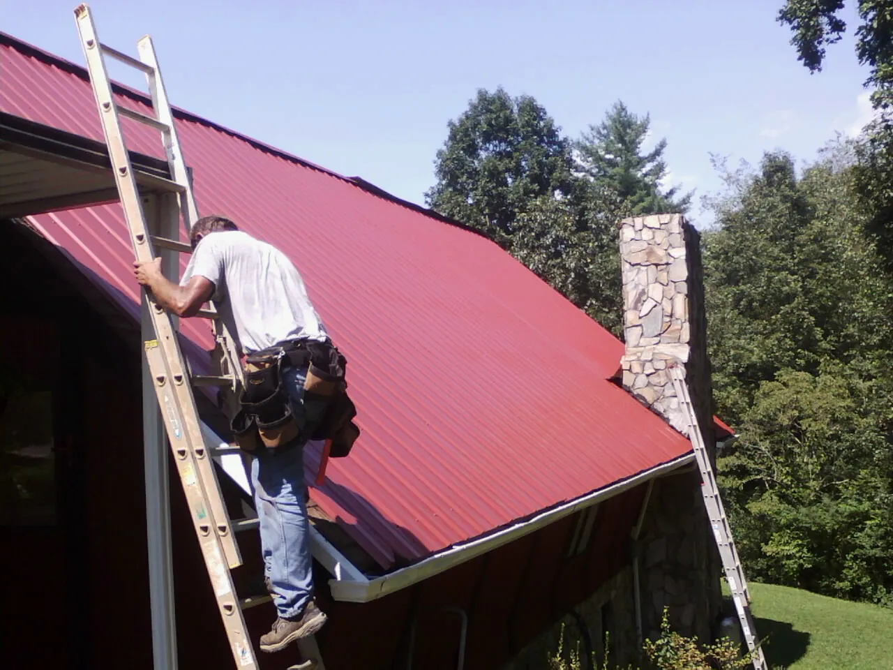 Roofer on a ladder beside a red metal roof and stone chimney — Soffit & Fascia Repair by Roofing Brothers in Whitestone